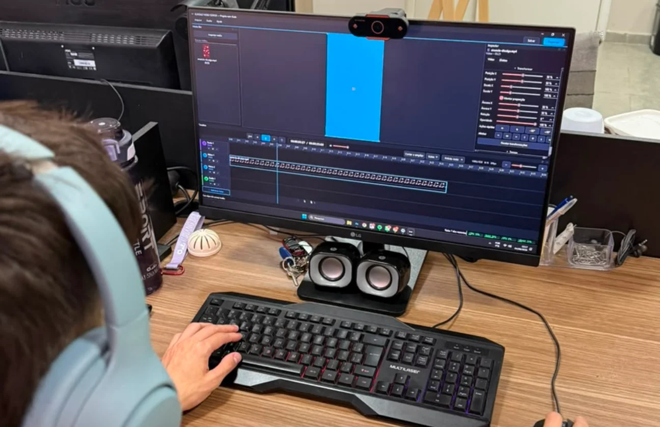 Pessoa de fones de ouvido editando no BLINDAO VIDEO EDITOR em um monitor de mesa, com a mão no teclado; na tela, a prévia em 9:16, o Inspetor à direita e a timeline com os clipes